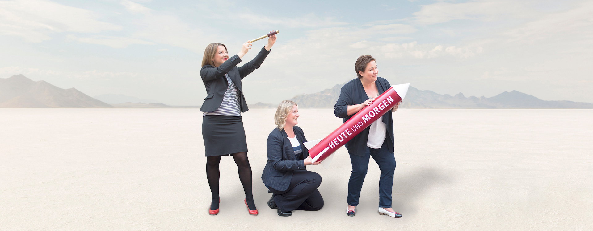 Drei Frauen in Anzügen in einer Wüste mit einer Rakete in ihren Händen, auf der "HEUTE UND MORGEN" steht